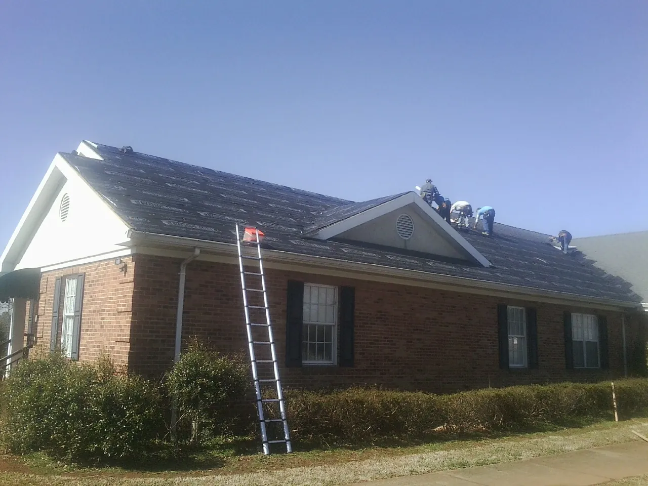 Crew tearing shingles off a brick ranch roof with a ladder set — Emergency Roof Repair work by our crew in Leslie